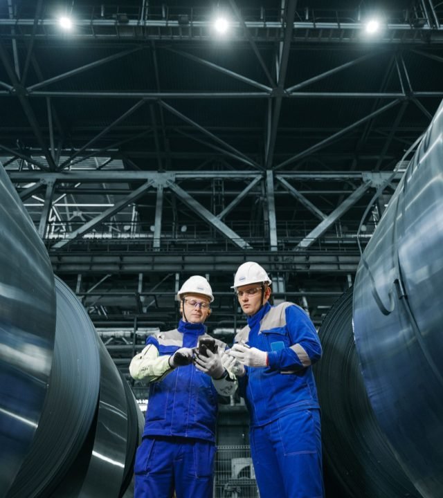 Two workers in protective gear examine steel coils in a modern industrial facility.