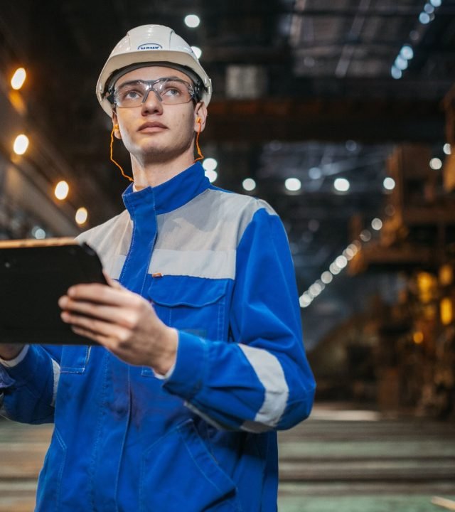 Engineer in safety gear uses a tablet in a large industrial factory setting, focused and diligent.